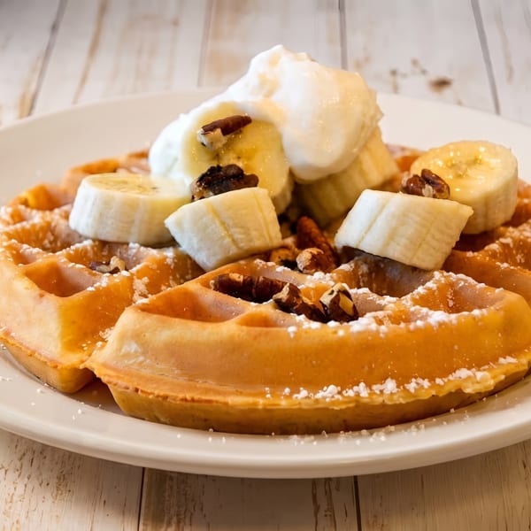 Golden waffle topped with sliced bananas, whipped cream, and pecans on a white plate.